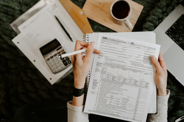 Pessoa conferindo planilhas e documentos financeiros ao lado de notebook e calculadora, imagem ilustrativa